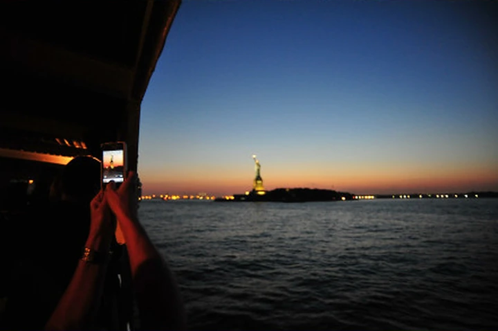 A passenger photographs the Statue of Liberty from a boat at sunset.