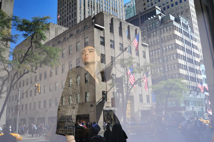 A mannequin in a shop window layered with reflections of New York buildings, trees and flags.